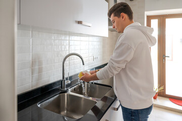 Caucasian brown-haired man in the kitchen. He is rinsing the dishes. In a white kitchen with black marble. There is natural light inside in Spain.