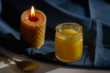 Fototapeta na wymiar Natural bee yellow honey in a glass jar with a wax candle in the shape of a honeycomb and a spoon with honey on a blue cloth