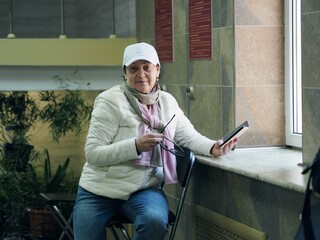 An elderly woman in a white jacket sits in the sanatorium hall near the window and uses a cell phone.