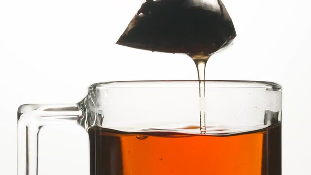 Tea Bag Over a Full Cup of Tea. The tea bag in the form of a pyramid is slowly removed from the mug and a thin stream of tea flows from it
