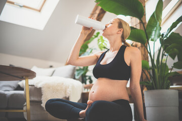 Fototapeta na wymiar Young happy and cheerful beautiful pregnant woman taking a break, hydrating, drinking water from the botle after home well being workout program.