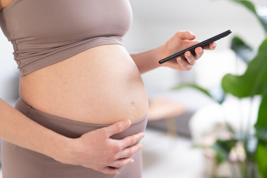 Close-up photo of pregnant female belly. Woman holding and using mobile smart phone application at home interiors. Pregnancy, technology, online shopping, preparation and expectation concept.