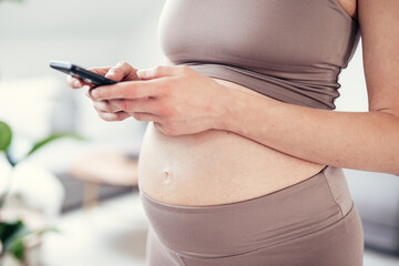 Close-up photo of pregnant female belly. Woman holding and using mobile smart phone application at home interiors. Pregnancy, technology, online shopping, preparation and expectation concept.