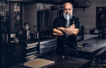 Professional elderly cooker smoking vape in kitchen of restaurant