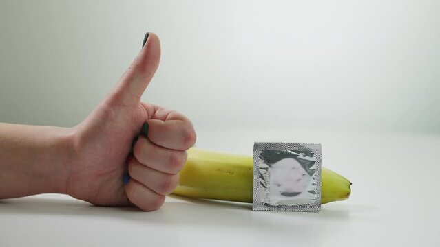 Close-up female hand gesturing thumb up with condom lying at banana. Unrecognizable Caucasian young woman advertising contraceptive at white background. Sex education
