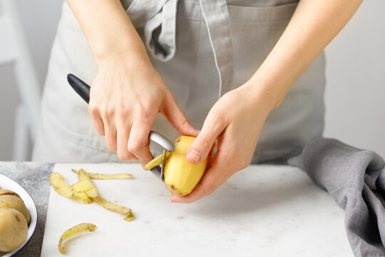 Woman in apron peeling potato on marble board. White background. Hands peeling potato with kitchen peeler.