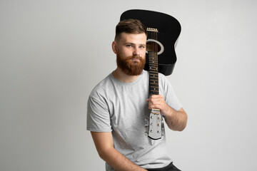 Handsome brunette bearded man musician in a grey t-shirt sitting on a chair and holding a acoustic guitar on a shoulder on a white background studio.