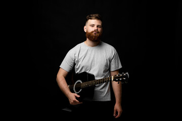 Handsome brunette bearded man musician standing and holding a acoustic guitar in a hand and looks in a camera on a black background studio. Ready to play a music.