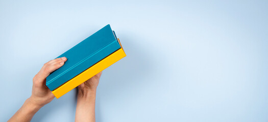 Female hands holding two blue and yellow color books over light blue background. Education,...