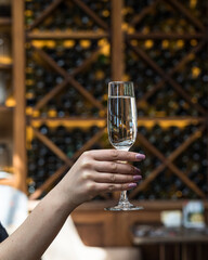 A woman's hand with a glass of white wine. In the background is the interior of the restaurant, a rack with wine bottles. Warm soft light.