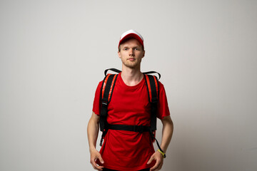Young courier in a red uniform t-shirt and with red food thermo bag on a shoulder standing isolated on white background studio. Food delivery service.