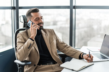 Businessman talking on a mobile phone and writing in a notebook while sitting in his office