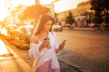 Fototapeta premium long-haired girl in glasses with phone, coffee and bag walks around the city at sunset