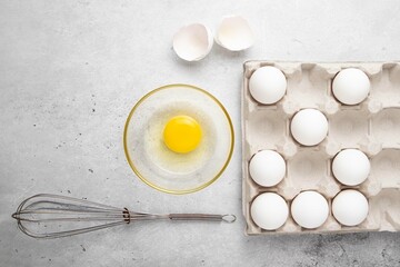 Fresh white organic chicken eggs in paper tray, broken egg and shell, whisk on light background. Baking background. Top view, copy space or empty place for text. Selective focus.