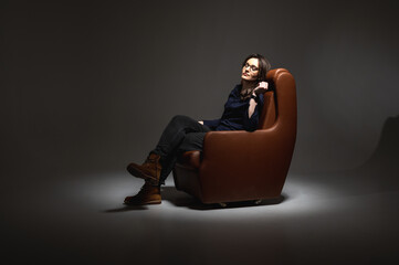 Beautiful, young, emotional, tired and frustrated woman sitting on a brown leather chair in a secluded isolated room, a beam of light illuminates. Low key. Hard days