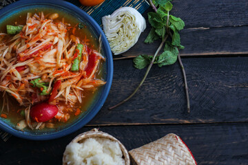 Spicy papaya salad served in a blue dish with sticky rice in a bucket and long beans, dried chilli and vegetables.