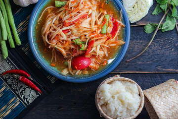 Spicy papaya salad served in a blue dish with sticky rice in a bucket and long beans, dried chilli and vegetables.