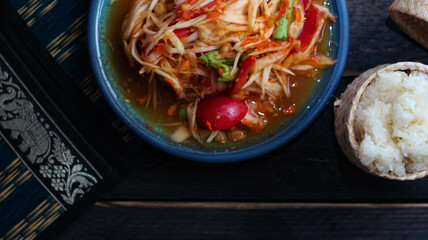 Spicy papaya salad served in a blue dish with sticky rice in a bucket and long beans, dried chilli and vegetables.