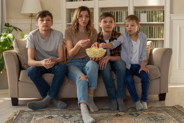 Happy family, a woman and three boys, are sitting on the sofa in the living room with a bowl of popcorn, watching TV