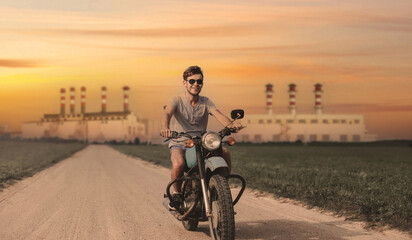 Fototapeta na wymiar a male motorcyclist rides an old retro motorcycle on a sandy road against the backdrop of an industrial city, the concept of gasoline transport and the oil refining industry.