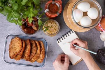 Overhead view of a woman hand writing in a spiral notebook the word MENU next to some glass jars full of meal.