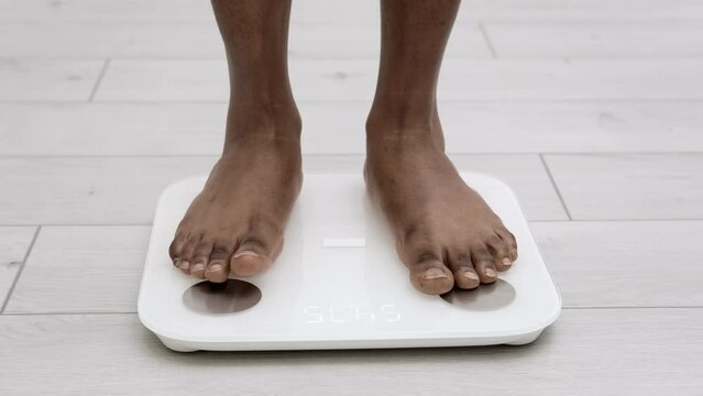 Legs of black girl standing on scale to measure weight. African American Female bare feet with weight scale at home. dieting, control and measuring 