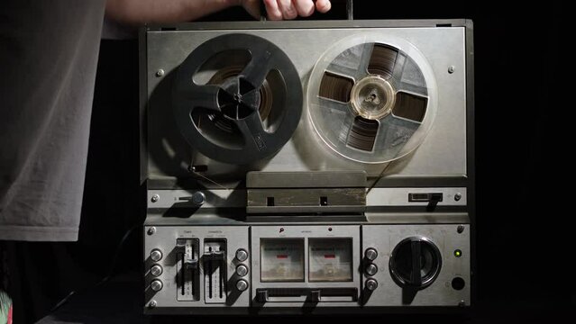man puts old vintage reel player on table and turns it on. carry gray retro magnetic tape player, put it on light area and press play. Inscriptions in Russian: volume, level, left, right, timbre