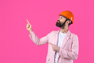 Portrait of young construction engineer wear orange hard hat, in a pink jacket standing on red studio background. The man points with his hand.