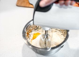 Bakery chef using her bakery equipment. Hand beater beating egg, sugar and flour in silver bowl on white background countertop for homemade dessert. A person mixing fresh ingredient for pastry at home
