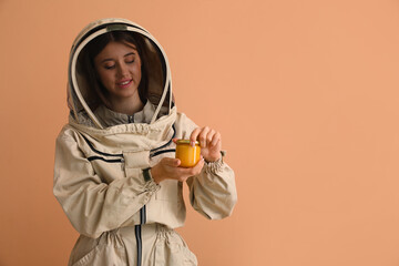 Female beekeeper in protective suit with jar of honey on beige background