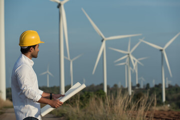 engineer wearing yellow hard hat standing with drawing against wind turbine.