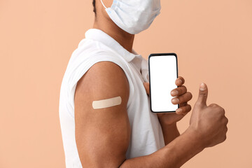 Young man in mask with applied medical patch and mobile phone showing thumb-up on beige background,...