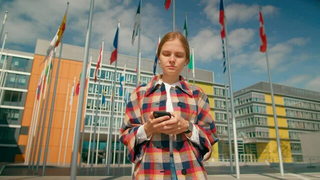 Young Woman Walks using Mobile app on Smart Phone in European Parliament administrative office Courtyard with Waving Flags of EU Countries in Luxembourg City. 4K tracking shot