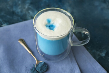 Glass cup of blue matcha tea and spoon with powder on blue grunge background