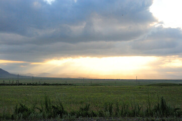 The bright rays of the setting sun shine through a huge cloud on the evening valley in the hilly steppe.