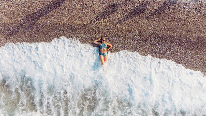 Aerial top view young woman in swimsuit lying on the stone beach, waves touch her legs. Young woman sunbathing and relaxing on the stone beach. Summer travel concept