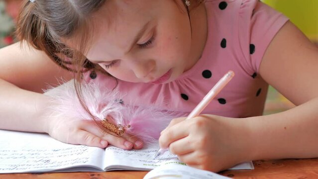 A little girl in a pink dress is studying lessons, a concentrated look at a notebook, incorrect posture, low head tilt, writes numbers with her left hand.