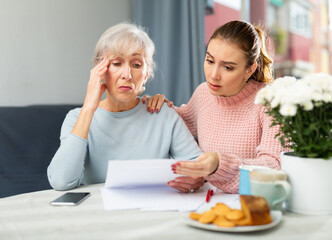 Portrait of elderly woman and her adult daughter looking at papers at home, worrying about debts on bills.