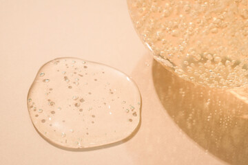 Drop of transparent cosmetic textured gel with bubbles and bottle with cosmetic liquid on beige background with copy space. Bath gel background, selective focus