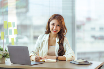 Charming Asian woman with a smile sitting and taking notes with computer laptop on her desk, enjoying work. Looking at camera.