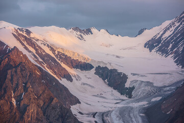 Dramatic aerial view to high snow mountain range in early morning at dawn. Awesome scenery with sunlit snow mountains in cloudy sky at sunrise. Scenic landscape with large glacier in sunrise colors.