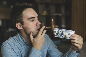 Young brutal businessman lighting cigar with 100 dollar bill as symbol of wealth and success. concept of wealth and extravagance. - obrazy, fototapety, plakaty