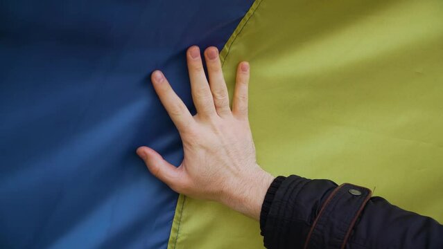 Close up hand of Ukrainian man patriot touches large developing flag of his country on tracked tank during war for independence and freedom. Crisis, peace, stop aggression in whole world