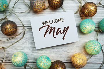 Welcome May text on paper card with LED cotton balls top view on wooden background