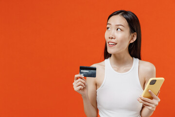 Fototapeta premium Vivid young woman of Asian ethnicity 20s in white tank top using mobile cell phone hold credit bank card order delivery booking tour looking aside isolated on plain orange background studio portrait.