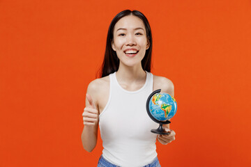 Overjoyed vivid excited young woman of Asian ethnicity 20s year old in white tank top hold in hands Earth world globe showing thumb up like gesture isolated on plain orange background studio portrait