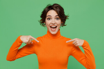 Young surprised shocked amazed woman 20s wear casual orange turtleneck point index fingers on herself isolated on plain pastel light green color background studio portrait. People lifestyle concept.