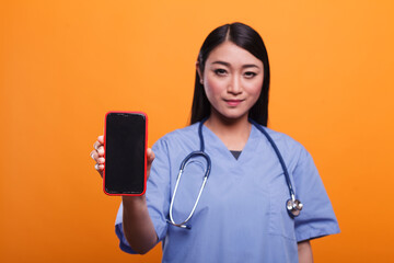Hospital caregiver wearing medical instrument while holding mobile cellphone on orange background....