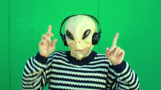 Alien dancing and listen music with headphones against a green wall background. Extraterrestrial with human clothes. Concept of victory and satisfaction. Happiness and freedom immigration concept