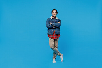 Full size body length young brunet bearded man 20s in denim jacket keep hands crossed look camera smiling isolated on plain pastel light blue background studio portrait. Winter cold season concept